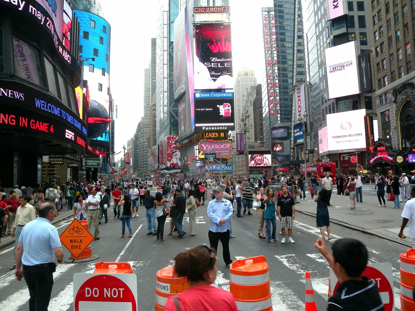 Color photograph of crowds walking in the roadway of Broadway at Times Square, with orange traffic barrels and signs closing the street to cars and illuminated billboards above.