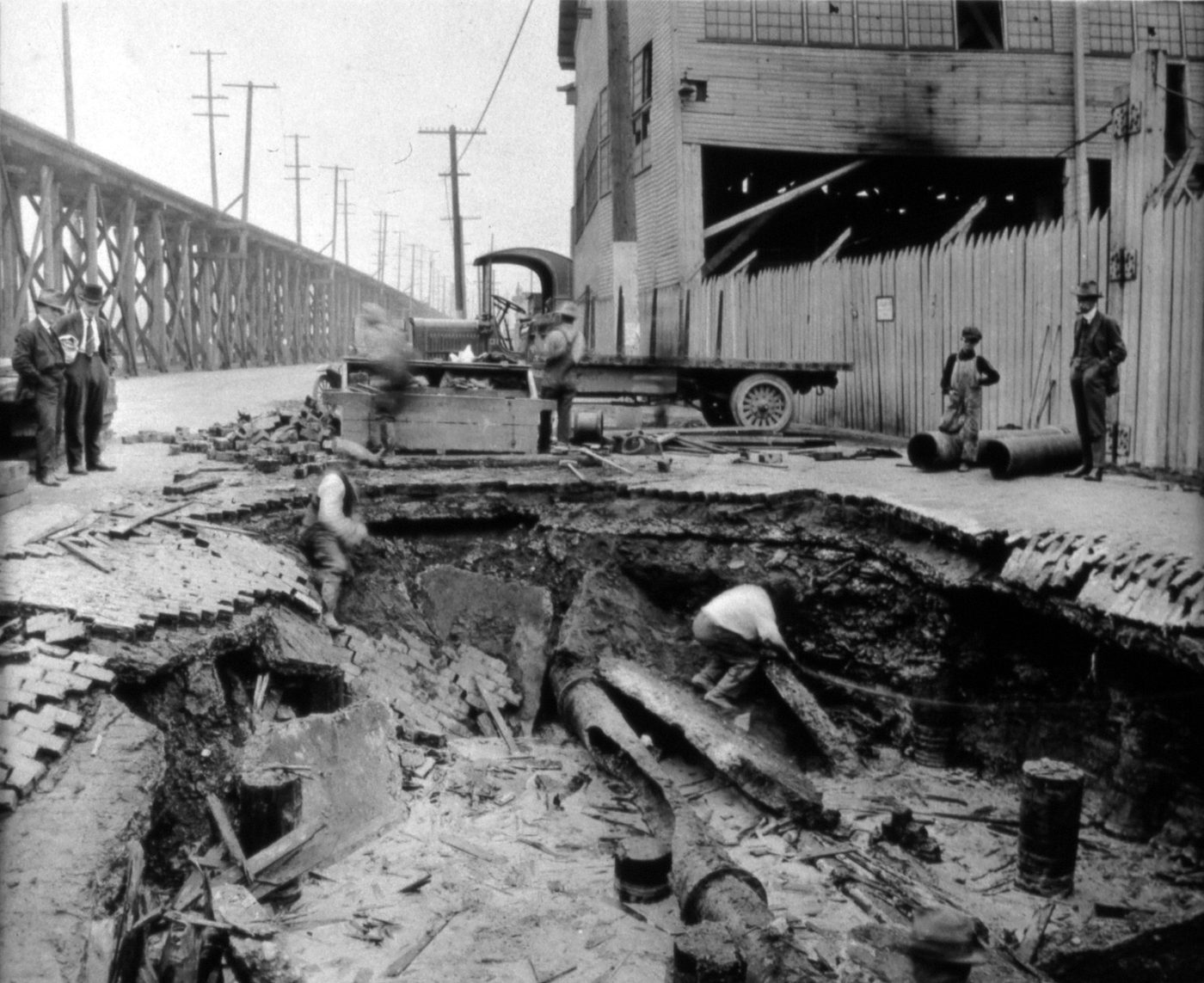 Sepia photograph of a deep trench dug across a city street, a worker in the pit beside an exposed pipe, men watching from above and a truck parked nearby.